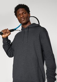Giovane uomo in felpa grigia scura con un racchetta da tennis nera e blu sulle spalle, in piedi contro uno sfondo chiaro e uniforme.