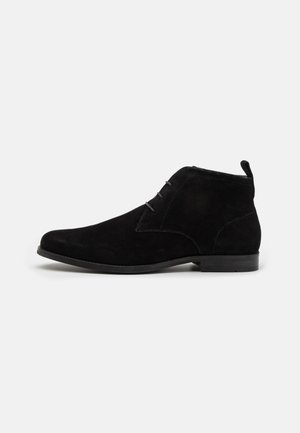 Schwarze Wildleder-Ankle-Boots mit abgerundetem Zeh, zwei Ösen für Schnürsenkel und einem kleinen Absatz. Mit einer Zuglasche an der Rückseite für einfaches Anziehen.
