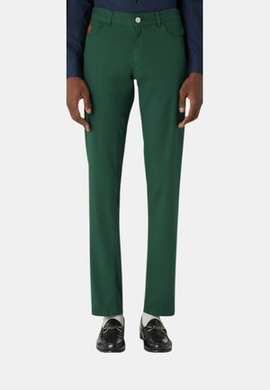 Uomo che indossa pantaloni verde scuro a gamba dritta, mocassini neri con catene e una camicia blu scuro, in piedi davanti a uno sfondo uniforme.