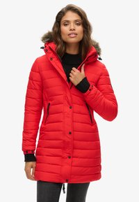 Giacca parka imbottita rossa con cappuccio foderato in pelliccia removibile, tasche con zip, dettagli neri e polsini a coste. Texture liscia e design di lunghezza media.