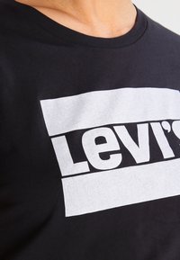 Černé bavlněné tričko s velkým bílým logem Levi's v obdélníkovém designu, s kulatým výstřihem a krátkými rukávy.