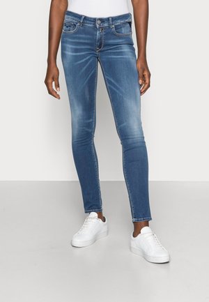 Persona con jeans ajustados azul claro con un ligero desgaste y zapatillas blancas, de pie frente a un fondo liso y claro.