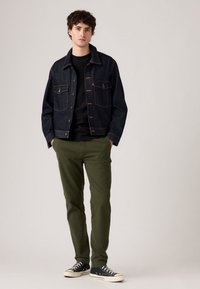 Young man wearing a dark denim jacket, black shirt, olive green pants, and black sneakers, standing with hands in pockets against a plain background.
