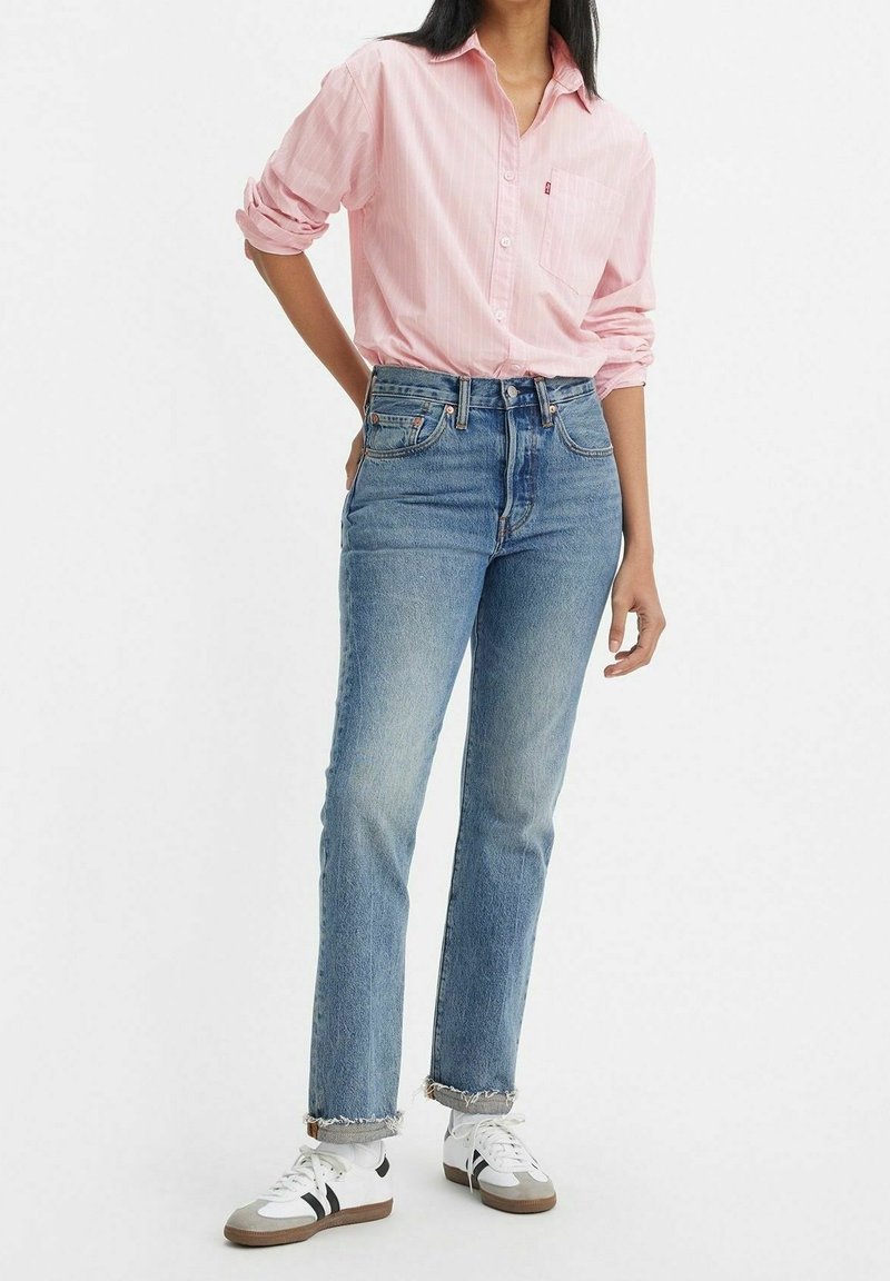 Personne portant une chemise rose clair à boutons, un jean bleu taille haute avec des ourlets bruts et des baskets blanches à rayures noires, debout avec une main derrière le dos.