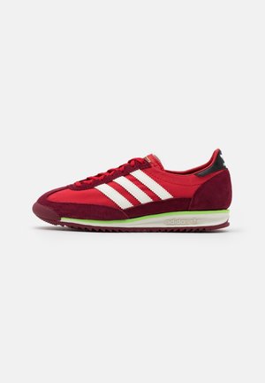 Baskets Adidas rouges avec bandes blanches, semelle à accents verts, dessus en daim et tissu, et languette de talon noire, vues de côté sur fond blanc.