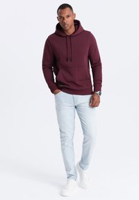 Felpa bordeaux con cordini, tasca a kangaroo e dettagli del logo discreti. Abbinata a jeans di denim azzurri chiari e scarpe da ginnastica bianche.