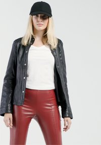 Schwarze Lederjacke, weißes V-Ausschnitt-T-Shirt und hochtaillierte rote Lederimitat-Hosen. Das Model trägt eine schwarze Mütze, die Hände an den Seiten. Glatte Texturen.