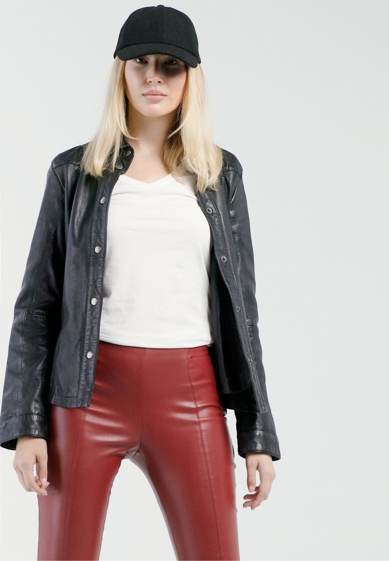 Schwarze Lederjacke, weißes V-Ausschnitt-T-Shirt und hochtaillierte rote Lederimitat-Hosen. Das Model trägt eine schwarze Mütze, die Hände an den Seiten. Glatte Texturen.
