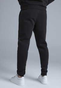 Musta puuvillasekoitteesta valmistetut collegehousut, joissa on kapeneva leikkaus, sivutaskut ja resorit lahkeensuissa, yhdistettynä valkoisiin tennareihin.