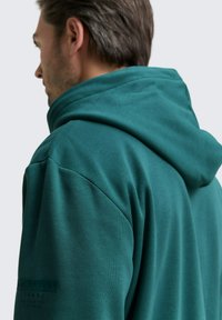 Mann mit kurzen braunen Haaren, der einen türkisfarbenen Kapuzenpullover trägt, von hinten und von der Seite gezeigt, mit sichtbaren Details zu Schultern und Kapuze.