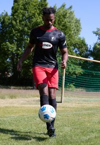 Tricou sportiv texturat negru cu accente roșii și logo, pantaloni scurți roșii, șosete negre și ghete negre. Lovind o minge de fotbal cu model alb și albastru pe iarbă.