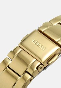 Kultainen metallinen kellonranneke kiillotetulla pinnalla, jossa on toisiinsa liittyviä linkkejä ja lukko, jossa on kohokuvioitu brändin nimi "GUESS."