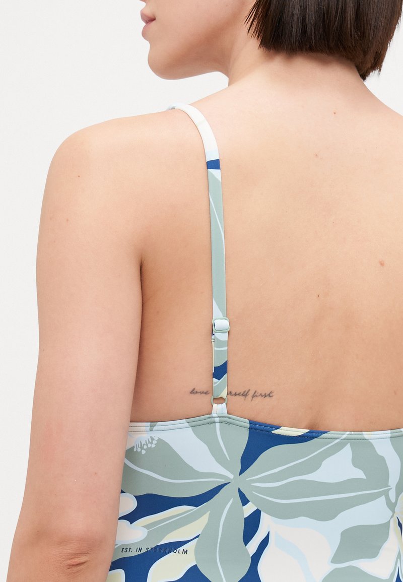 Einteiliger Badeanzug mit floralem Muster in Blau und Grün, mit verstellbaren Trägern und einem niedrigen Rückendesign. Enthält ein sichtbares Tattoo.