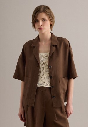 Jong persoon met kort lichtbruin haar draagt een bruin kort mouw jasje met knopen over een wit gehaakt topje en bijpassende bruine broek.