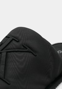 Sandal cu toc plat negru, cu un superior texturat, având un logo proeminent și un design structurat. Vârf rotunjit și material moale, cu finisaj mat.