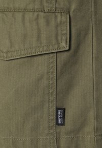 Olivgrüner Stoff mit Fischgrätenmuster, ausgestattet mit einer Klappentasche und einem kleinen Markenetikett an der Seite. Sichtbare Nahtdetails.