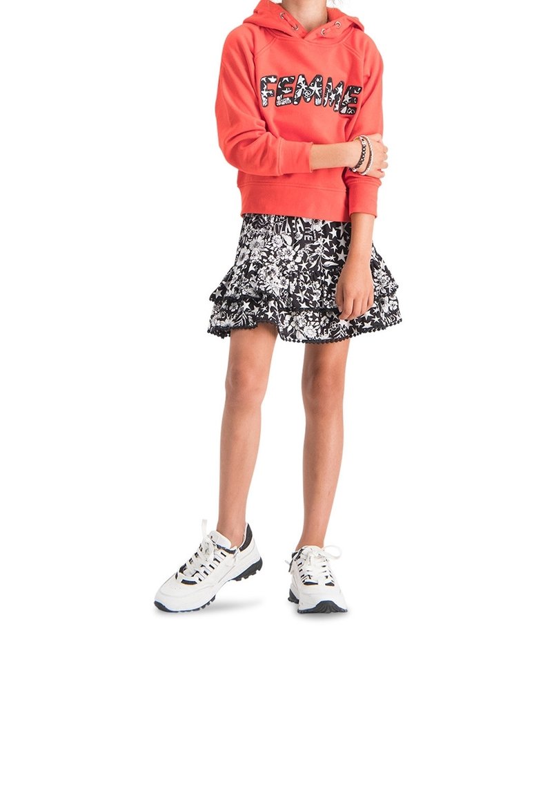 Punainen huppari, jossa on "FEMME" -grafiikka, kerrostettu mustavalkoinen kukkakuvioinen röyhelöhame ja massiiviset valkoiset lenkkarit mustilla yksityiskohdilla.