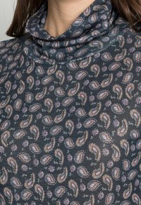 Långärmad polokrage i mörk tyg, med ett paisley- och blommönster i nyanser av rosa och ljusblått på en texturerad bakgrund.