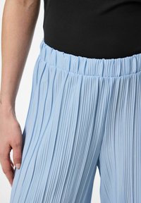 Ljusblå veckade byxor med elastisk midja, tillverkade av textil med struktur, närbild som visar midjan och veckdetaljer.