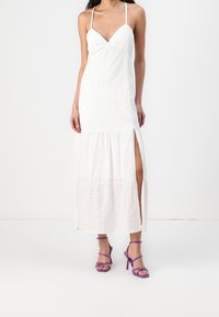 Vestido largo blanco con cuerpo entallado y falda de capas. Presenta tirantes finos tipo espagueti y una abertura lateral alta hasta el muslo. Se combina con tacones morados con tiras.