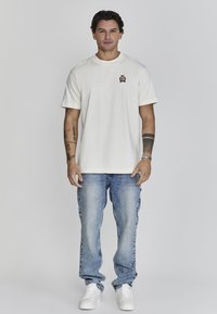 Camiseta de manga corta color crema con cuello redondo y pequeño logotipo bordado, combinada con jeans de mezclilla azules y zapatillas blancas.