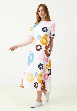 Kvinde iført en hvid midi-kjole med store farverige blomsterprint i blå, pink, orange, brun og gul, kombineret med hvide sneakers.