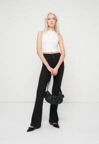 Svarte vide jeans og en hvit ermeløs topp. Modellen holder en liten svart håndveske med teksturert finish og har på seg svarte spisse flate sko.