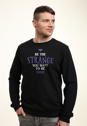 Man met kort haar en oorringen, gekleed in een zwarte sweatshirt met paarse tekst "Wees de vreemde die je wilt zijn", kijkt naar rechts met zijn handen in zijn zakken.