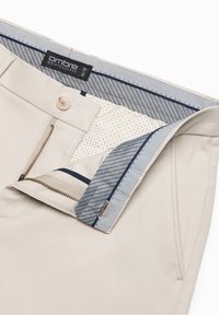 Pantaloni beige con cerniera e bottone parzialmente aperti, che mostrano la fodera interna della vita a righe e motivi, con etichetta del marchio sfumata.