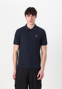 Jūras zilas krāsas polo krekls ar klasisko apkakli, īsām piedurknēm, trīs pogu kārtojumu un mazu dzeltenu logotipu uz krūšu. Gluda auduma tekstūra.
