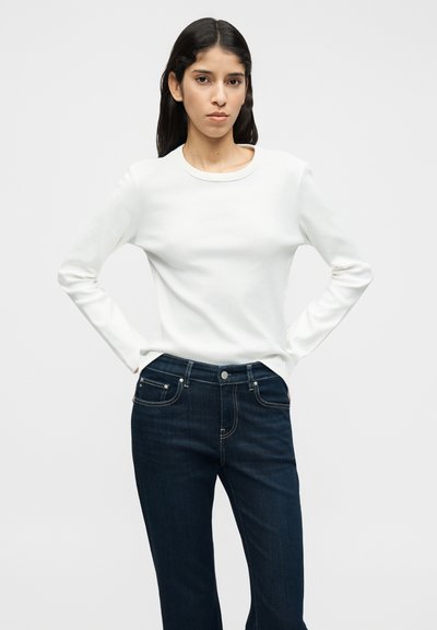 Λευκή μπλούζα με μακριά μανίκια και στρογγυλή λαιμόκοψη, συνδυασμένη με σκούρο μπλε τζιν παντελόνι. Το ύφασμα φαίνεται λείο, με εφαρμοστή γραμμή και απλό σχέδιο.