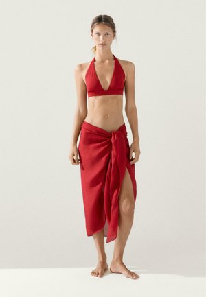 Žena stojící naboso, oblečená v červeném halter bikini topu a odpovídající červené zavinovací sukni s předním uzlem a rozparkem.