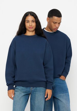 Deux personnes portant des sweats bleu foncé assortis et des jeans bleu clair se tiennent devant un fond blanc uni.