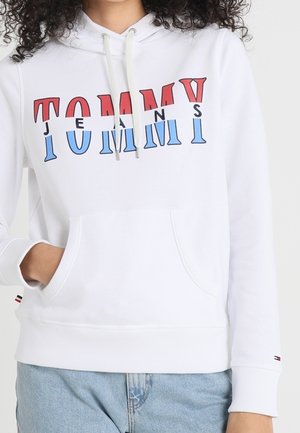Kobieta w białej bluzie z kapturem z czerwonym i niebieskim napisem "TOMMY JEANS" i jasnoniebieskich dżinsach, ręka w kieszeni bluzy.