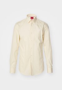 Chemise crème à manches longues boutonnée avec col pointu et étiquette intérieure rouge avec le nom de la marque "HUGO" sur fond uni.