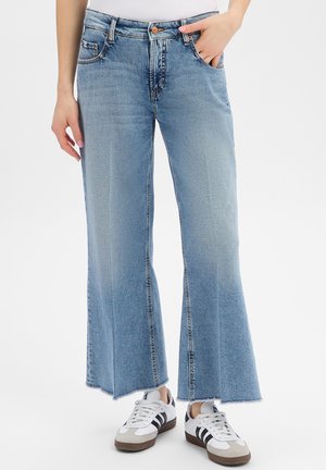 Hellblaue, hochtaillierte Jeans mit weitem Bein und rohem Saum, kombiniert mit weißen Sneakers mit schwarzen Streifen, eine Hand in der Tasche, die andere locker entspannt.