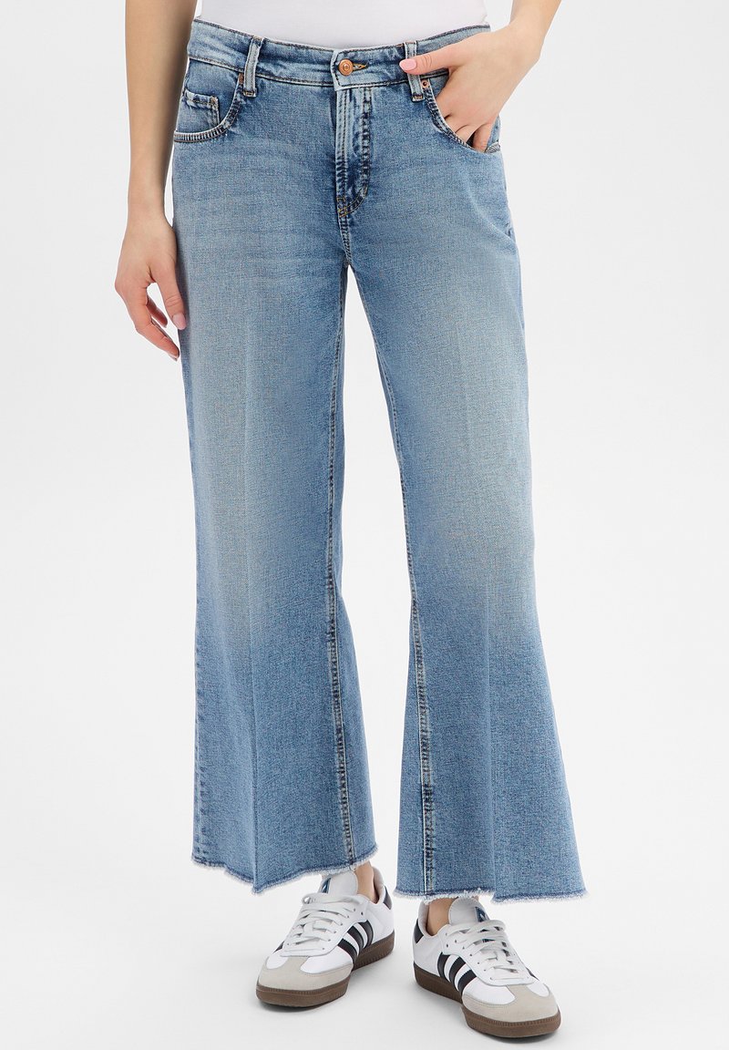 Hellblaue, hochtaillierte Jeans mit weitem Bein und rohem Saum, kombiniert mit weißen Sneakers mit schwarzen Streifen, eine Hand in der Tasche, die andere locker entspannt.