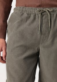 Pantalones cortos de canalé gris con cintura elástica y cordón. Bolsillos laterales y una textura suave. Vista en primer plano desde la cintura hacia abajo.