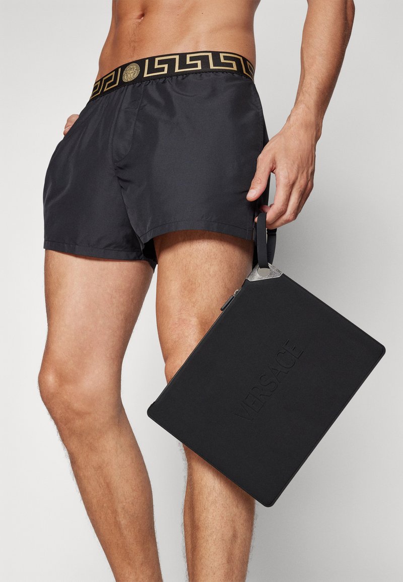 Pantaloni da bagno neri con una vita a motivo dorato, tenuti con una mano, affianco a una borsa nera rettangolare con la scritta "VERSACE" in rilievo.