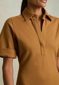 Kastanjebruine polo met een kraag, korte opgerolde mouwen en een verticale placket met een X-stiksel. Soepele stof.