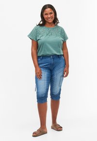 Yhdeksän vihreä puuvillapaita, jossa on kuvioitu yläosa, yhdistettynä vaaleansiniset denim capri-housut, joissa on sivuvetoketju ja frilla helmassa.
