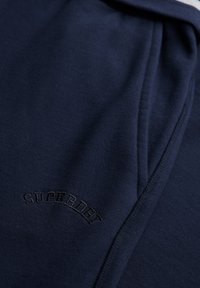 Tissu bleu marine avec une couture détaillée et "SUPERDRY" brodé en fil sombre assorti près d'une couture.
