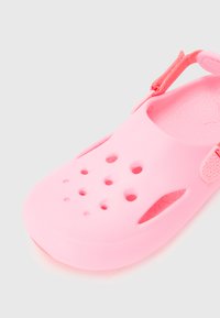 Roza gumijasta sandal s zaprtim nosom, z ventilacijskimi odprtinami in prilagodljivim trakom. Ima teksturiran vložek za stopala in gladek, fleksibilen material.