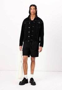 Schwarze Jeansjacke mit silbernen Knöpfen, kombiniert mit schwarzen Shorts und schwarzen Schnürschuhen. Das Modell hält eine beige Tote-Bag. Weißer Hintergrund.