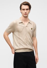 Μπεζ μπλούζα πόλο με κοντά μανίκια και V-λαιμόκοψη, με υφαντά σχέδια και μικρό κίτρινο λογότυπο πεταλούδας στο στήθος. Ύφασμα από λεία πλεκτή ύλη.