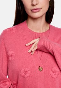 Rosa gestrickte Strickjacke mit floralen Applikationen, einem V-Ausschnitt, gerippten Bündchen und einem dekorativen Knopf. Getragen über einem passenden Oberteil.