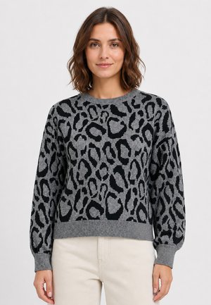 Woman wearing a gray and black leopard print sweater with long sleeves and light-colored pants against a plain background.