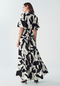 Maxi haljina u crnoj i bijeloj boji s apstraktnim uzorcima listova. Ima kratke rukave, struk s remenom i dizajn s višeslojnim suknjama.