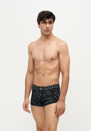 Hombre musculoso con cabello rizado oscuro lleva calzoncillos tipo bóxer negros con estampado y cintura negra sobre un fondo blanco liso.