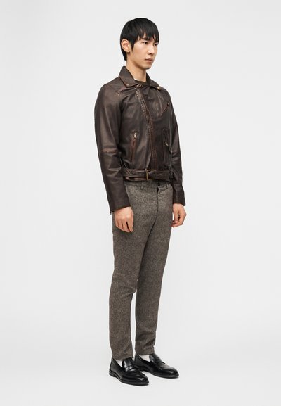 Νεαρός άνδρας στέκεται φορώντας ένα σκούρο καφέ δερμάτινο μπουφάν, γκρι μάλλινες παντελόνες, λευκές κάλτσες και μαύρα δερμάτινα loafers μπροστά σε μια απλή φόντο.